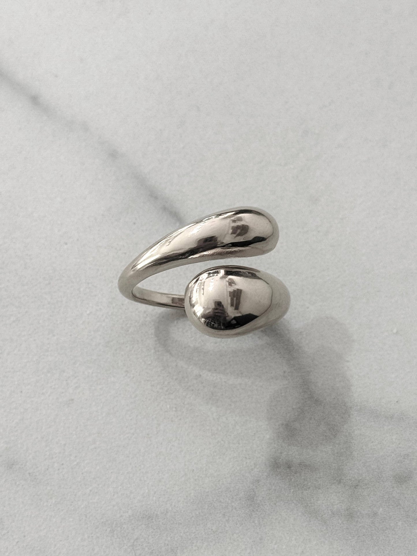 Bague en acier inoxydable de couleur argentée avec un anneau ouvert et croisé aux extrémités lisses et épaisses en forme de gouttes d'eau au fini poli miroir, présentée sur un fond en marbre blanc.