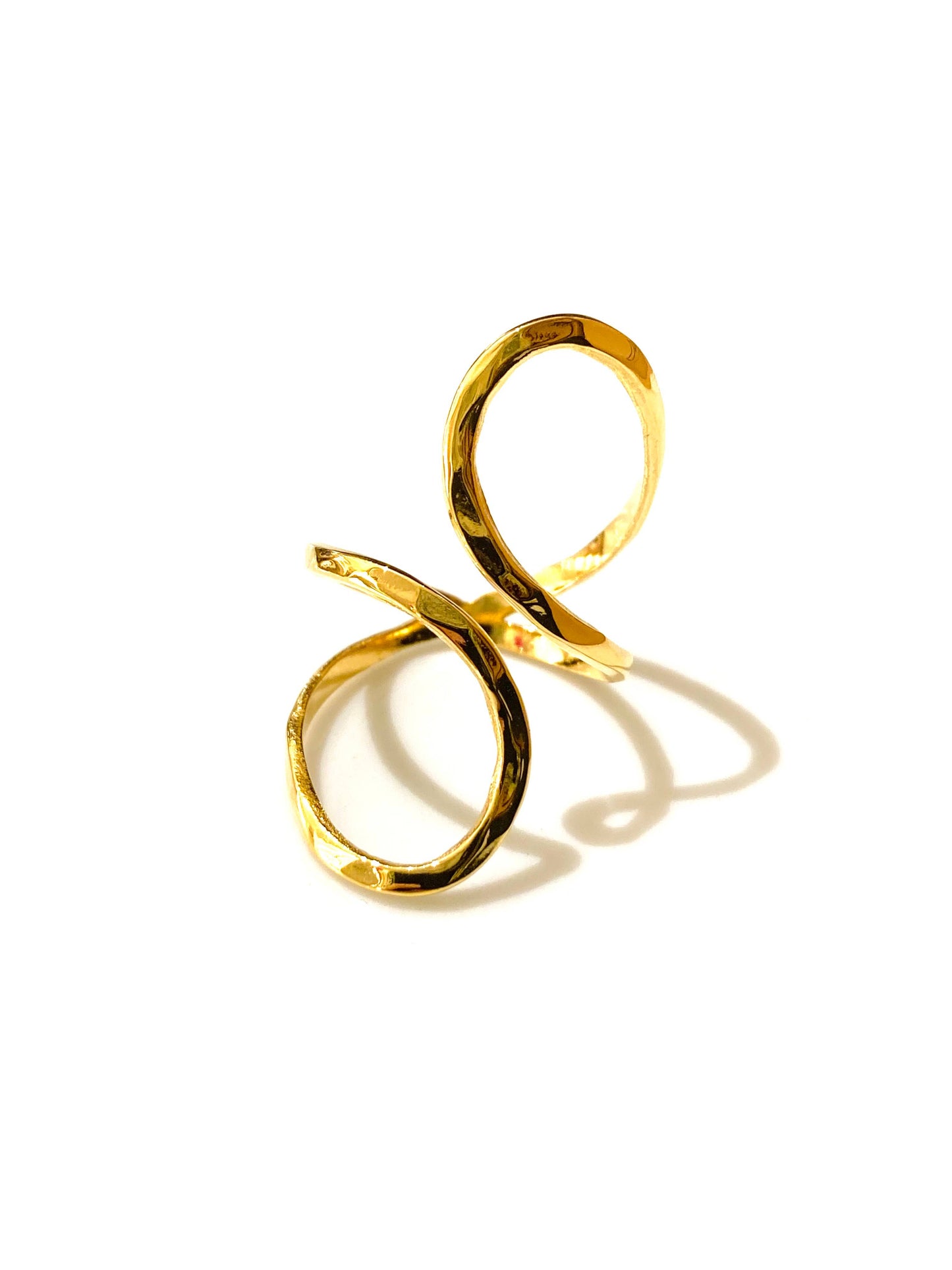 Bague ajustable en acier inoxydable doré, formée d'un ruban métallique plat qui s'enroule en spirale asymétrique avec un fini texturé martelé, photographiée de trois-quarts sur fond blanc avec une ombre portée marquée.