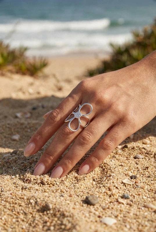 Bague statement en forme de fleur évidée en acier argenté avec strass centraux, portée au majeur d'une femme touchant le sable fin de la plage.