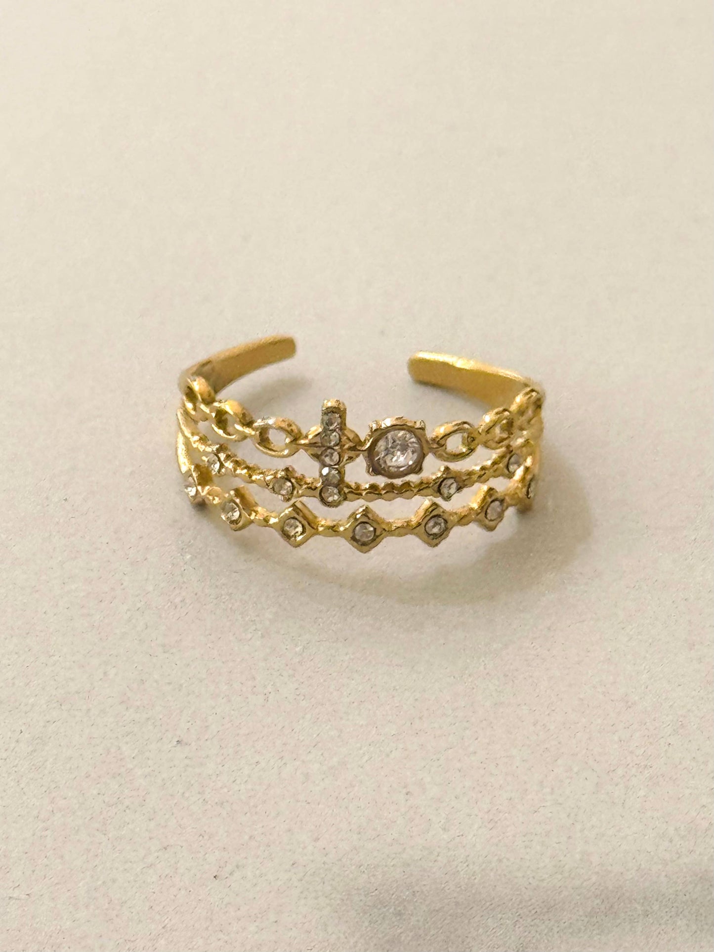 Bague multi-rangs ajustable en acier inoxydable doré, composée de trois anneaux texturés avec une petite croix pavée de strass, un petit solitaire rond et des motifs losanges, photographiée de face sur un fond blanc épuré.