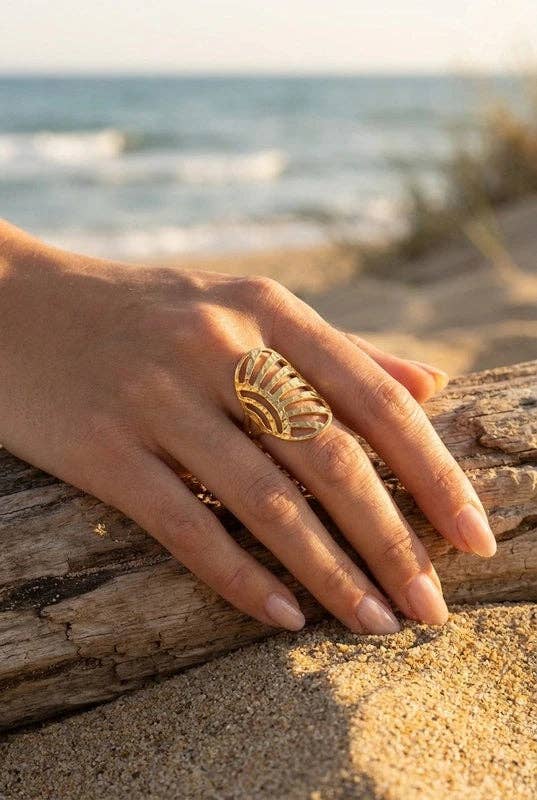 Bague large et allongée en acier inoxydable doré de forme éventail ciselé, portée à la main d'une femme posée sur un tronc de bois flotté en bord de mer.