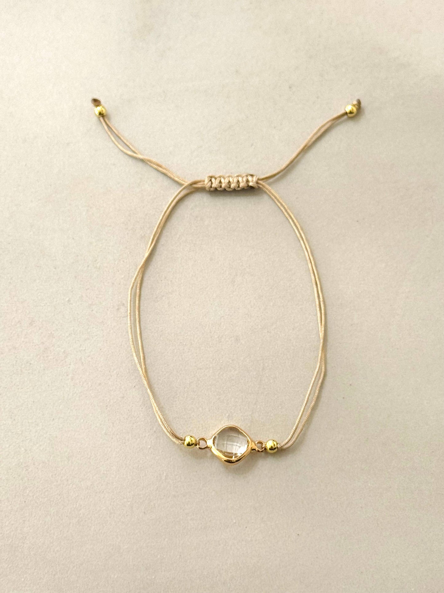Bracelet composé d'un double cordon beige réglable par un nœud coulissant, orné d'un intercalaire central en forme de losange arrondi en acier inoxydable doré entourant un cristal transparent facetté, posé à plat sur un fond beige clair.