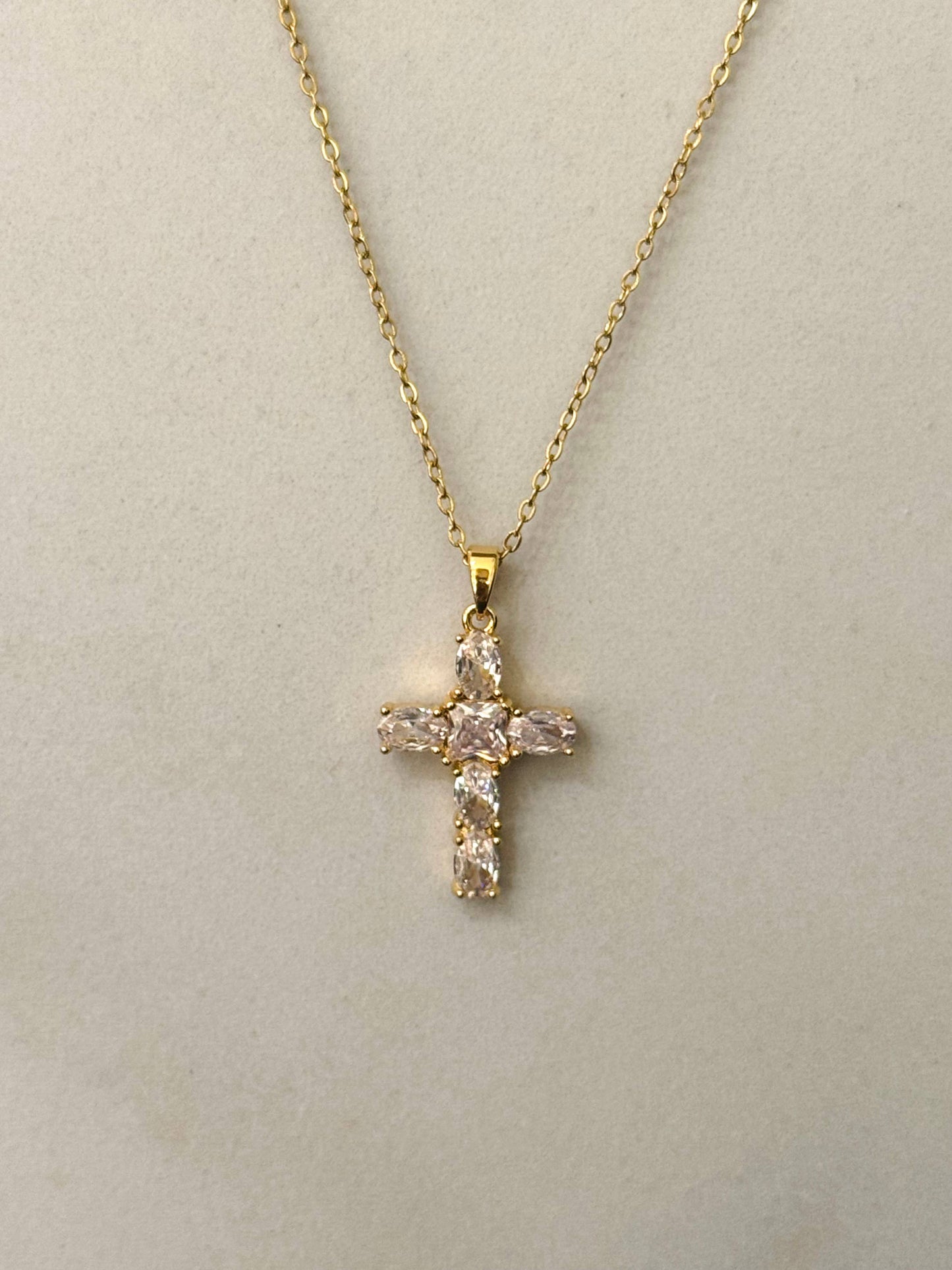 Collier à fine chaîne dorée en acier inoxydable avec un pendentif en forme de croix sertie d'oxydes de zirconium transparents et scintillants, présenté de face sur un fond beige clair épuré.