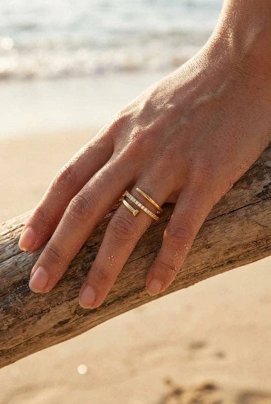 Bague ajustable en acier inoxydable doré portée au doigt sur une plage, dotée d'un design géométrique superposé orné d'une rangée de cristaux scintillants.