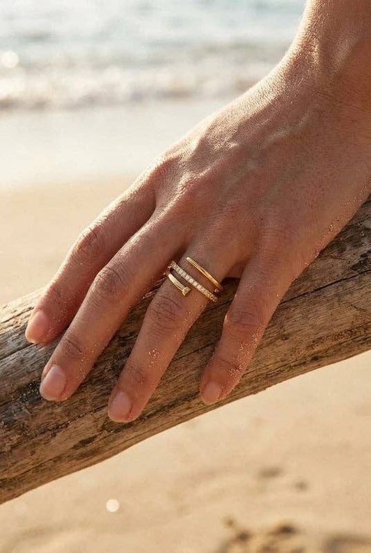 Bague ajustable en acier inoxydable doré portée au doigt sur une plage, dotée d'un design géométrique superposé orné d'une rangée de cristaux scintillants.