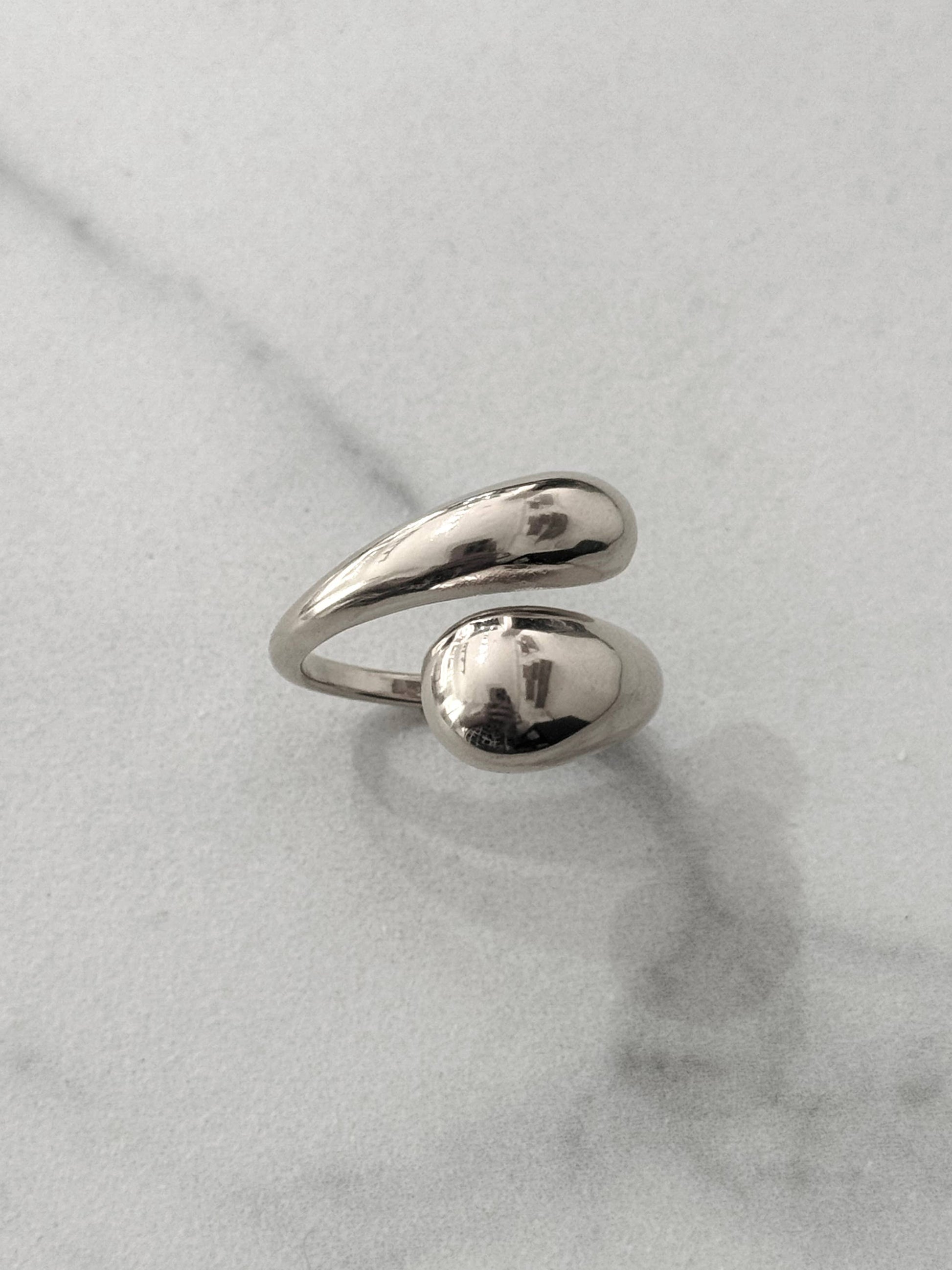 Bague en acier inoxydable de couleur argentée avec un anneau ouvert et croisé aux extrémités lisses et épaisses en forme de gouttes d'eau au fini poli miroir, présentée sur un fond en marbre blanc.