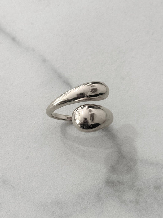 Bague en acier inoxydable de couleur argentée avec un anneau ouvert et croisé aux extrémités lisses et épaisses en forme de gouttes d'eau au fini poli miroir, présentée sur un fond en marbre blanc.