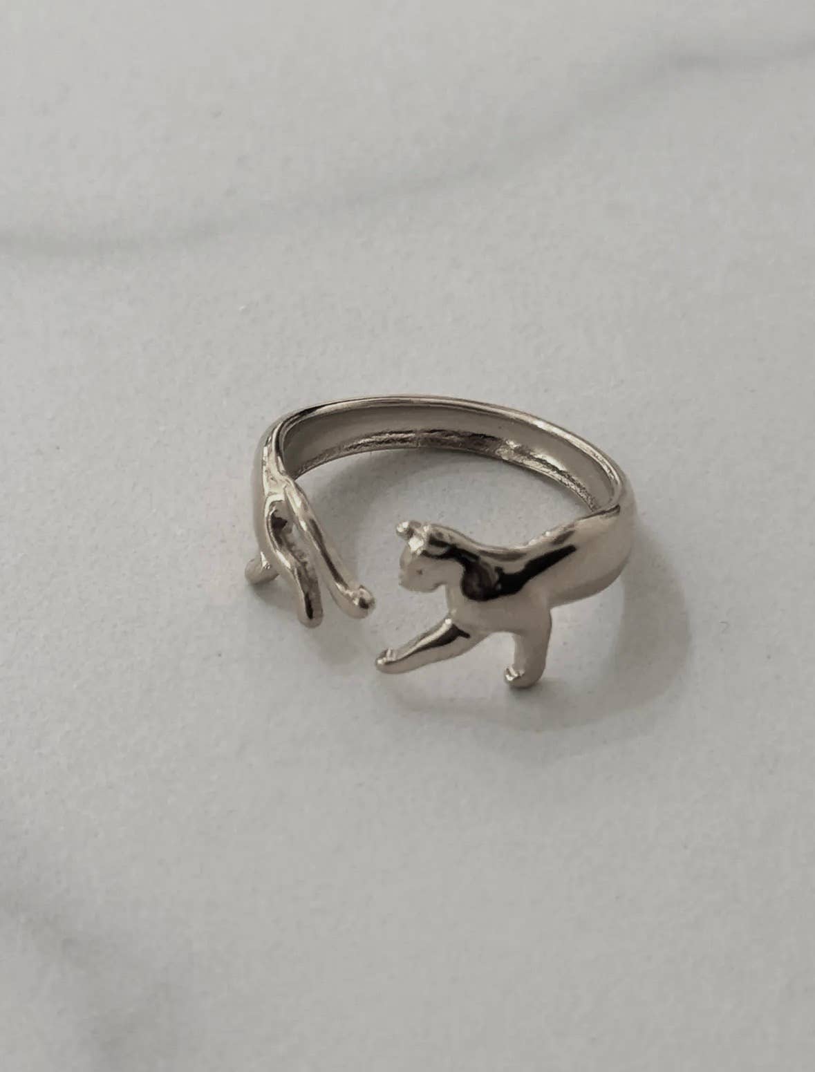 Bague ajustable ouverte en acier inoxydable argenté, sculptée de façon à représenter un petit chat brillant dont le corps enveloppe le doigt, avec d'un côté sa tête et ses pattes avant et de l'autre ses pattes arrière et sa queue, posée sur fond blanc.