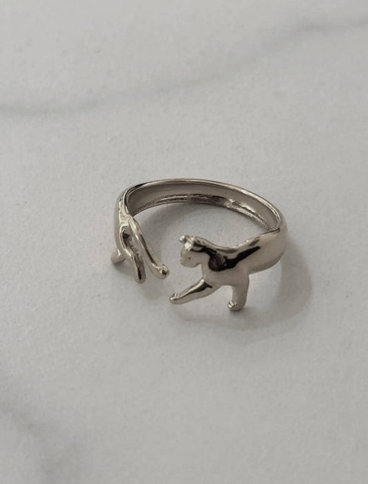Bague ajustable ouverte en acier inoxydable argenté, sculptée de façon à représenter un petit chat brillant dont le corps enveloppe le doigt, avec d'un côté sa tête et ses pattes avant et de l'autre ses pattes arrière et sa queue, posée sur fond blanc.