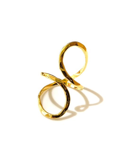 Bague ajustable en acier inoxydable doré, formée d'un ruban métallique plat qui s'enroule en spirale asymétrique avec un fini texturé martelé, photographiée de trois-quarts sur fond blanc avec une ombre portée marquée.