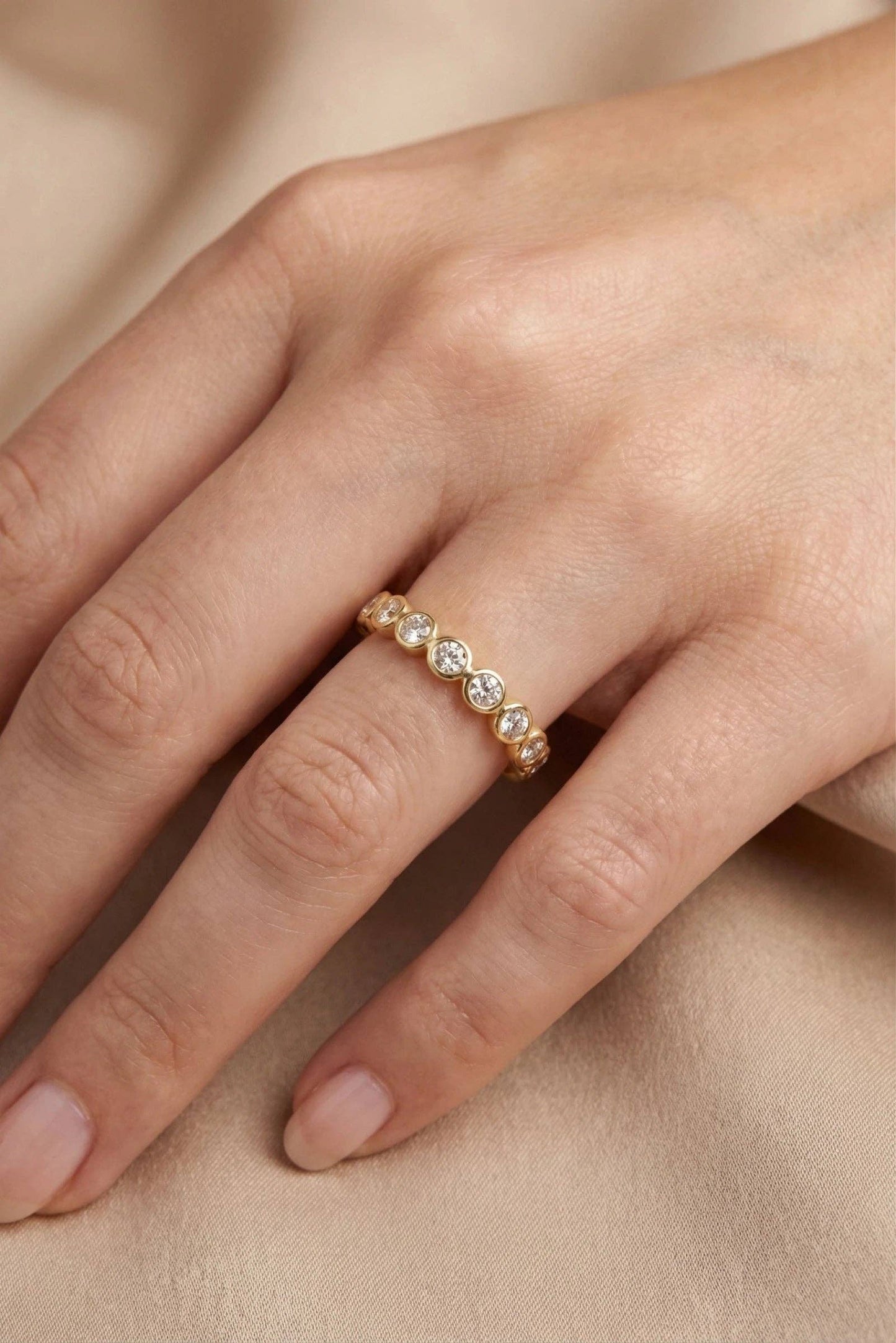 Bague de style éternité fine en acier doré avec alignement de petits strass ronds étincelants, portée au doigt d'une femme sur fond de tissu drapé beige beige.