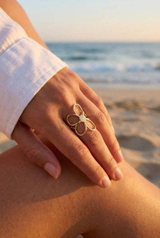Grande bague fleur ajourée en acier inoxydable doré avec cœur serti de brillants, portée à la main d'une femme en chemise blanche sur fond de plage au coucher du soleil.