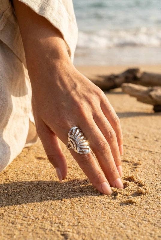 Bague statement allongée en acier inoxydable argenté avec des motifs de rayons ajourés, portée au majeur d'une femme posant sa main sur le sable d'une plage ensoleillée.
