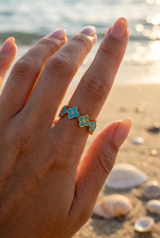 Bague anneau en acier inoxydable doré ornée d'une succession de trèfles émaillés bleu turquoise, portée au doigt d'une femme sur fond de plage ensoleillée avec des coquillages.