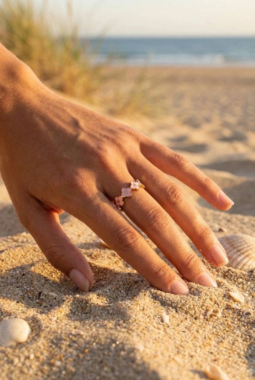 Bague en acier inoxydable doré composée d'une suite de petits trèfles à quatre pétales en émail rose clair, portée à la main posée sur le sable fin d'une plage.