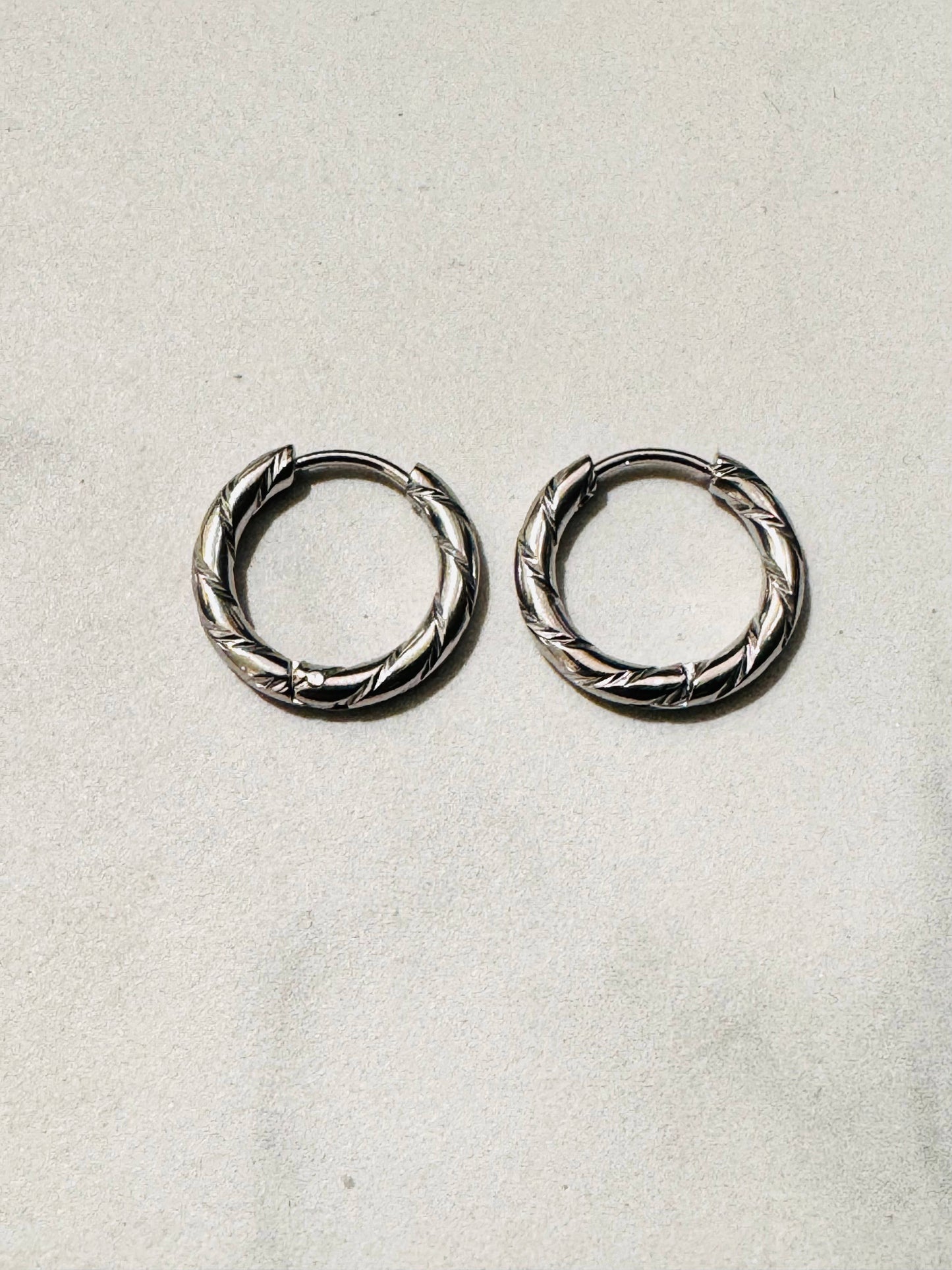 Paire de boucles d'oreilles créoles de type anneaux en acier inoxydable argenté, dotée d'un design texturé avec des gravures ciselées et polies, présentée de face sur un fond beige clair épuré.