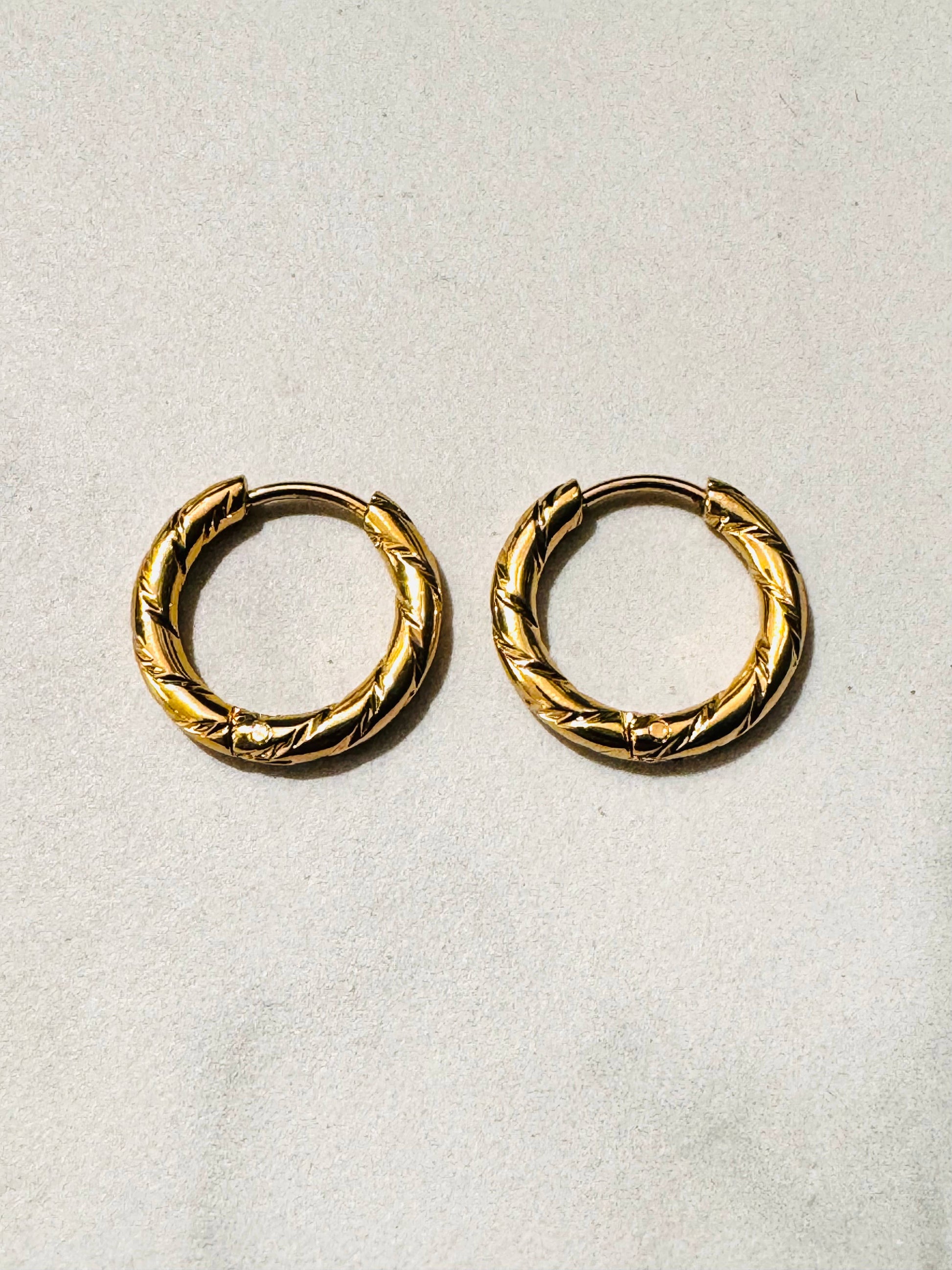 Paire de boucles d'oreilles créoles de type anneaux en acier inoxydable doré, présentant une surface épaisse ornée de motifs ciselés en oblique au fini miroir brillant, posée à plat sur un fond texturé beige clair.