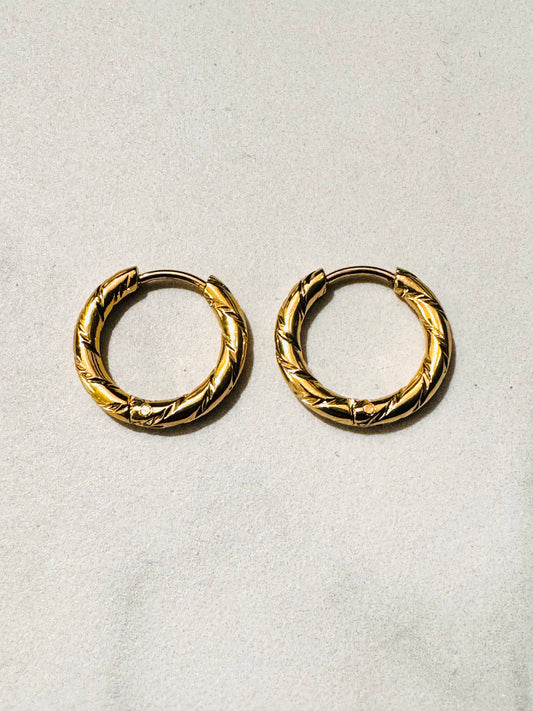 Paire de boucles d'oreilles créoles de type anneaux en acier inoxydable doré, présentant une surface épaisse ornée de motifs ciselés en oblique au fini miroir brillant, posée à plat sur un fond texturé beige clair.