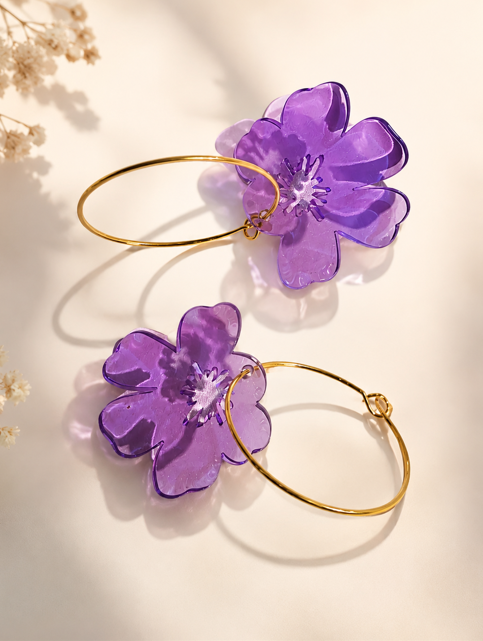 Paire de boucles d'oreilles créoles dorées ornées d'une grande fleur en résine translucide violette, posées sur un fond clair lumineux.