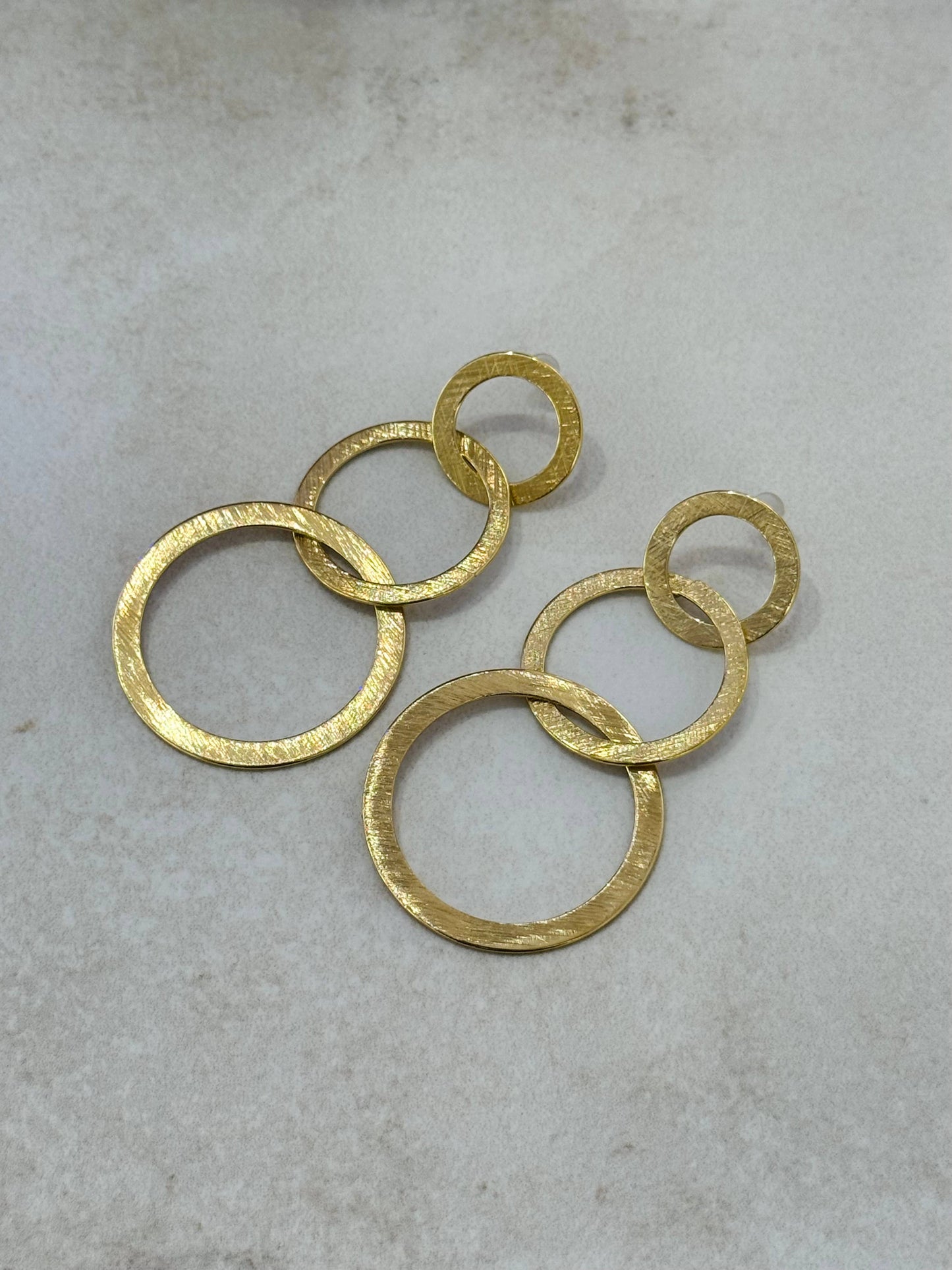 Paire de boucles d'oreilles pendantes en acier inoxydable doré, composées de trois cercles plats et imbriqués de taille croissante avec une texture d'or brossé, présentées à plat sur un fond beige clair texturé.