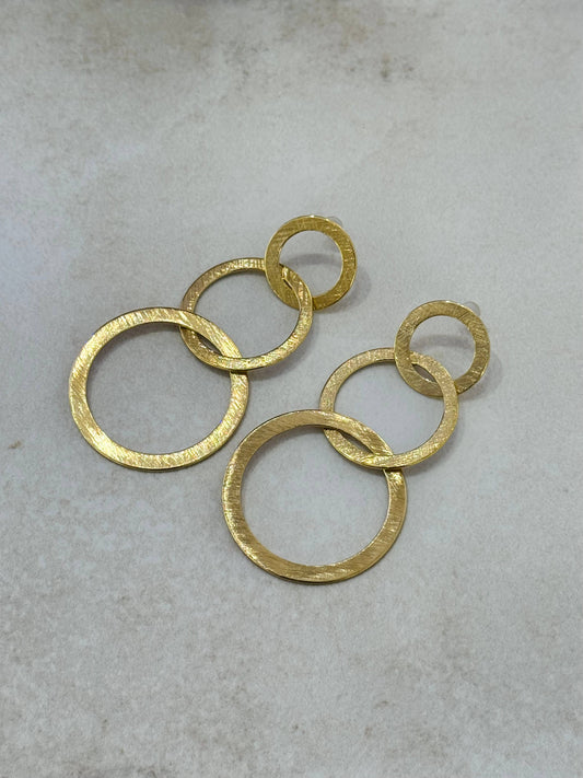 Paire de boucles d'oreilles pendantes en acier inoxydable doré, composées de trois cercles plats et imbriqués de taille croissante avec une texture d'or brossé, présentées à plat sur un fond beige clair texturé.