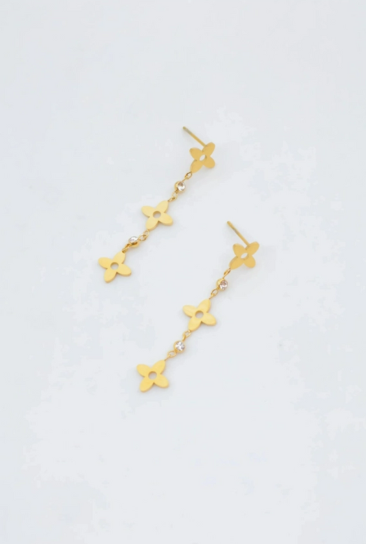 Paire de boucles d'oreilles pendantes de style clou en acier inoxydable doré, composées d'une chaîne alternant trois fleurs à quatre pétales ajourées et des petits strass brillants.