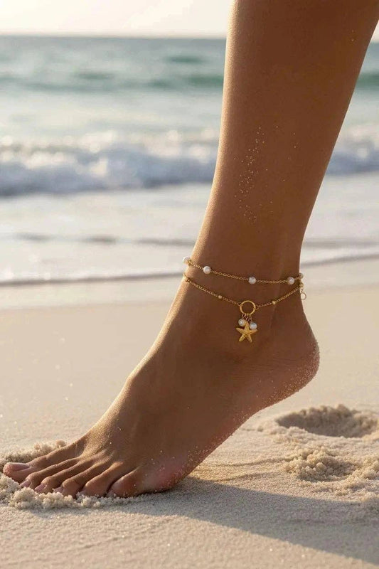 Bracelet de cheville double rang en acier inoxydable doré présenté sur un support de cheville blanc. Le premier rang est orné de perles blanches nacrées et le second d'une chaîne à billes avec trois charms suspendus : une étoile de mer ciselée, une petite perle et un strass brillant.
