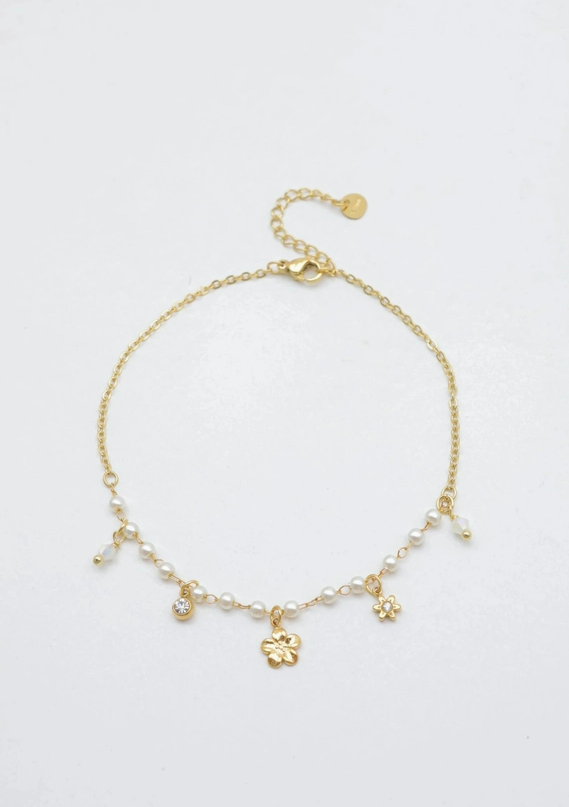 Bracelet de cheville en acier inoxydable doré posé à plat, composé d'une fine chaîne avec une rangée de perles blanches nacrées et des charms suspendus : une fleur à 5 pétales, une petite fleur étoile, un strass brillant et des pampilles transparentes.