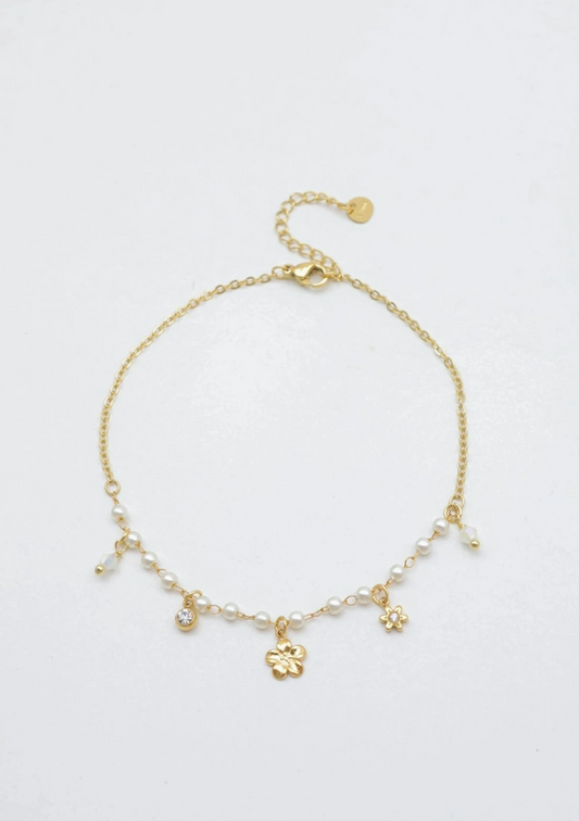 Bracelet de cheville en acier inoxydable doré posé à plat, composé d'une fine chaîne avec une rangée de perles blanches nacrées et des charms suspendus : une fleur à 5 pétales, une petite fleur étoile, un strass brillant et des pampilles transparentes.
