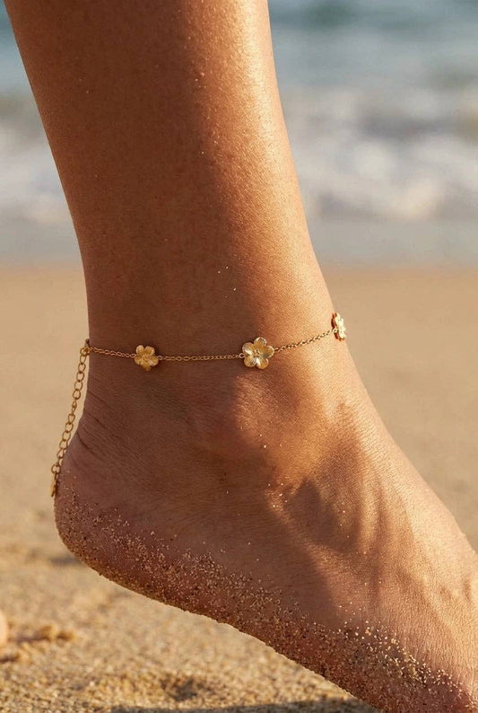 Bracelet de cheville en acier inoxydable doré orné de petites fleurs exotiques en relief, porté au pied sur une plage de sable fin en bord de mer.