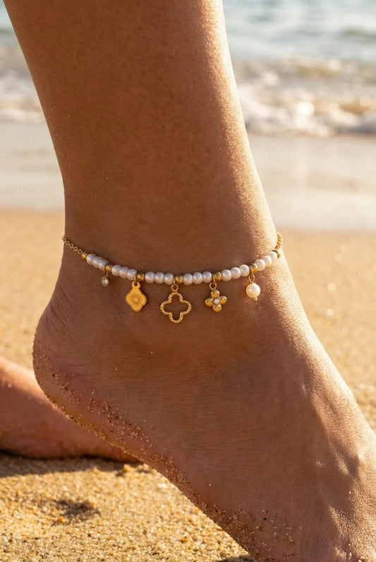 Bracelet de cheville en acier inoxydable doré et perles blanches nacrées, porté au pied sur une plage de sable fin. Le bijou est orné de 5 charms suspendus dont un grand trèfle évidé et une médaille ciselée.