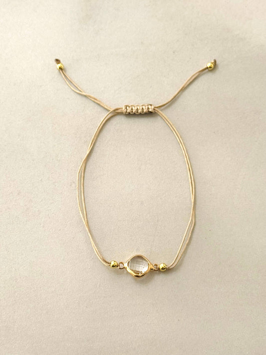 Bracelet composé d'un double cordon beige réglable par un nœud coulissant, orné d'un intercalaire central en forme de losange arrondi en acier inoxydable doré entourant un cristal transparent facetté, posé à plat sur un fond beige clair.