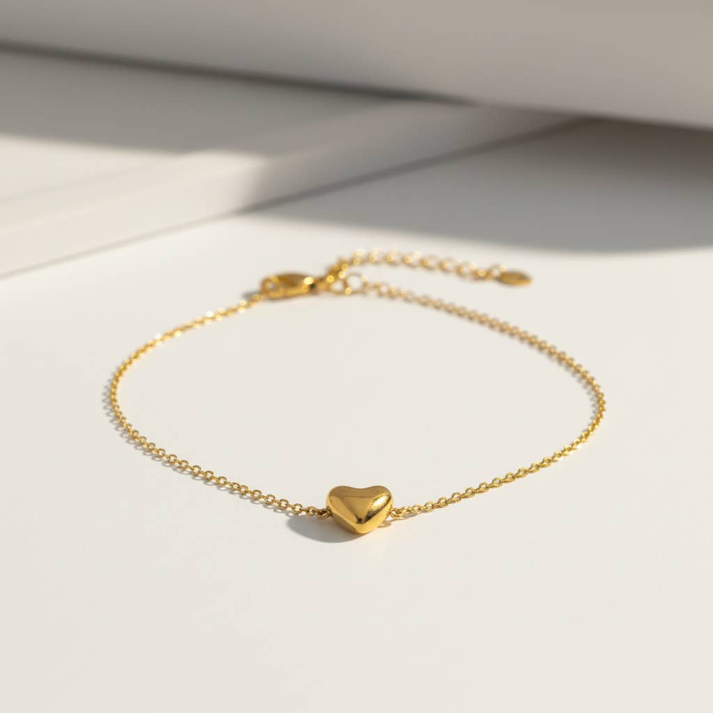Bracelet délicat composé d'une fine chaîne en acier inoxydable doré ornée d'un petit pendentif central en forme de cœur bombé en relief tridimensionnel au fini poli miroir brillant, posé sur une surface blanche lumineuse.