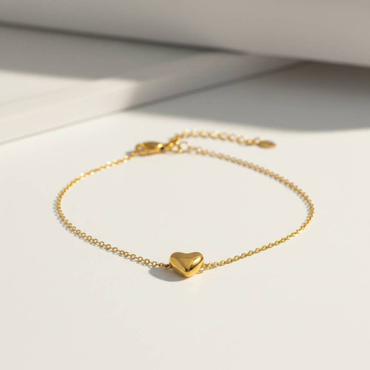 Bracelet délicat composé d'une fine chaîne en acier inoxydable doré ornée d'un petit pendentif central en forme de cœur bombé en relief tridimensionnel au fini poli miroir brillant, posé sur une surface blanche lumineuse.