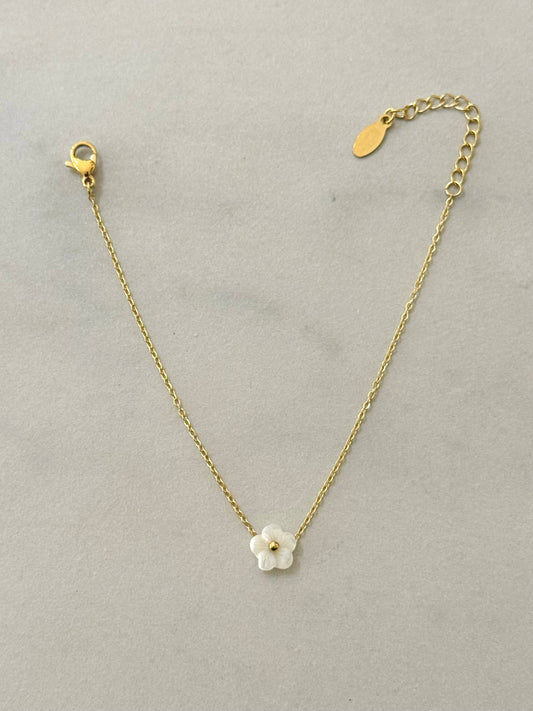Bracelet fin en acier inoxydable doré composé d'une chaîne en maille forçat, d'un fermoir mousqueton, d'une chaînette d'extension et d'une petite fleur centrale blanche aux reflets nacrés avec un cœur perlé doré, posé à plat sur fond gris clair texturé.