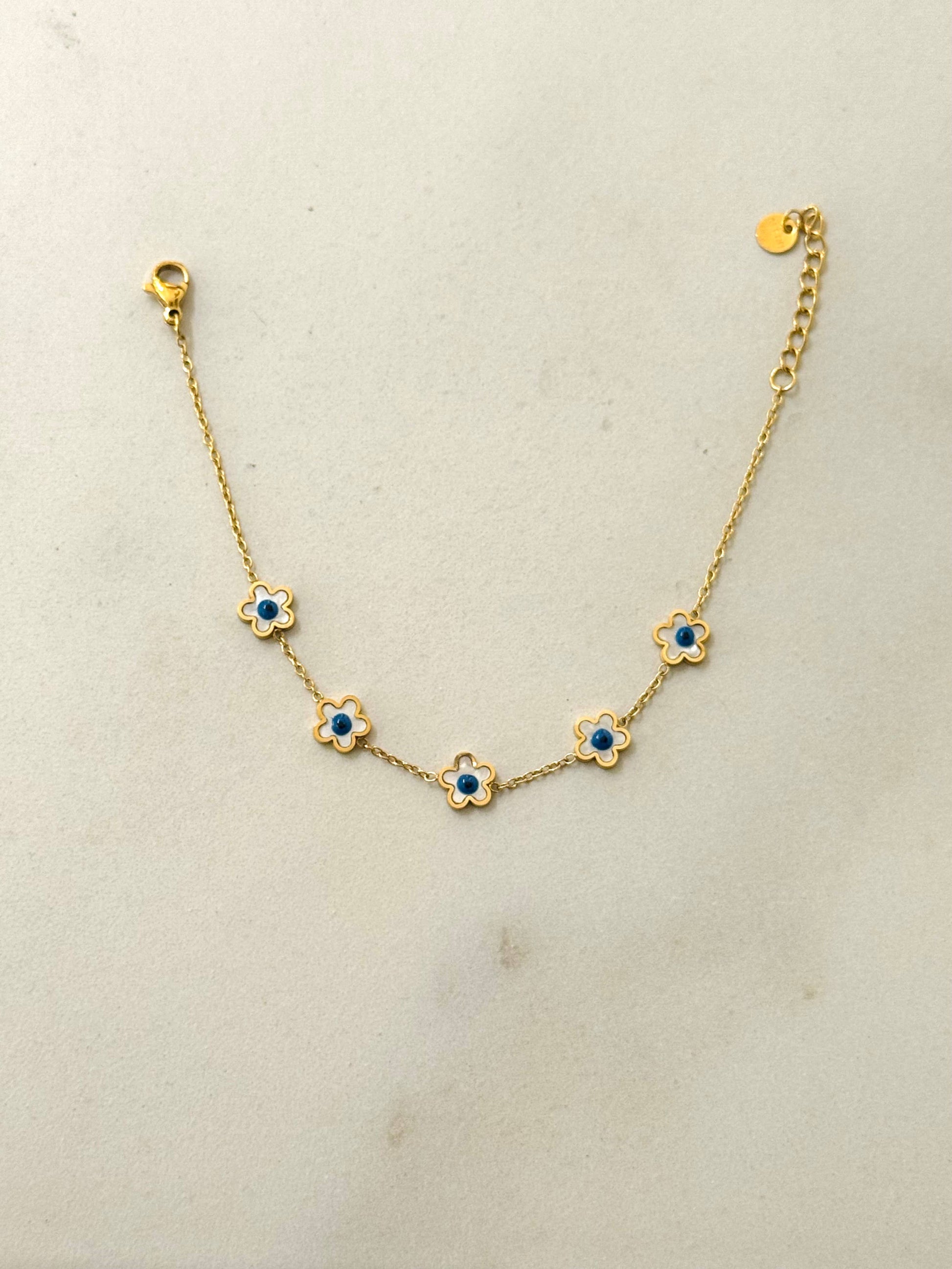 Bracelet fin en acier inoxydable doré composé d'une chaîne en maille forçat ornée de cinq petites fleurs aux pétales émaillés blancs et cœurs bleu roi, avec fermoir mousqueton et chaînette réglable, posé à plat sur fond gris clair.