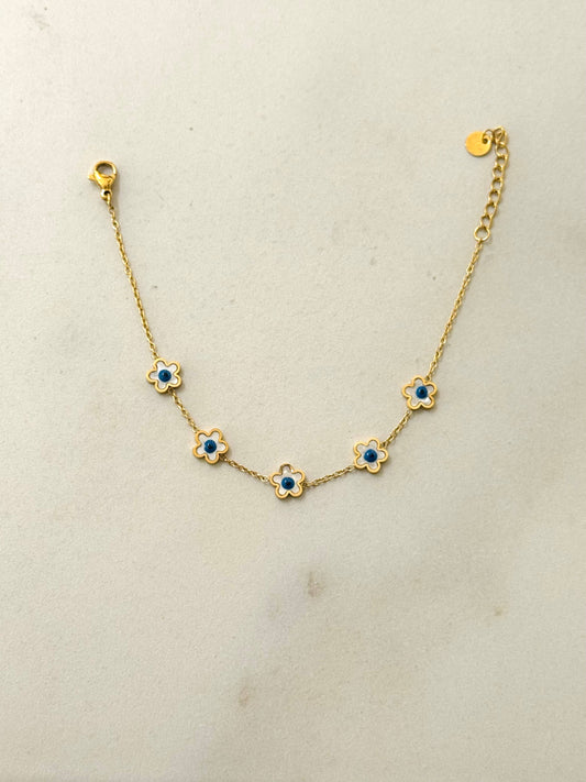 Bracelet fin en acier inoxydable doré composé d'une chaîne en maille forçat ornée de cinq petites fleurs aux pétales émaillés blancs et cœurs bleu roi, avec fermoir mousqueton et chaînette réglable, posé à plat sur fond gris clair.