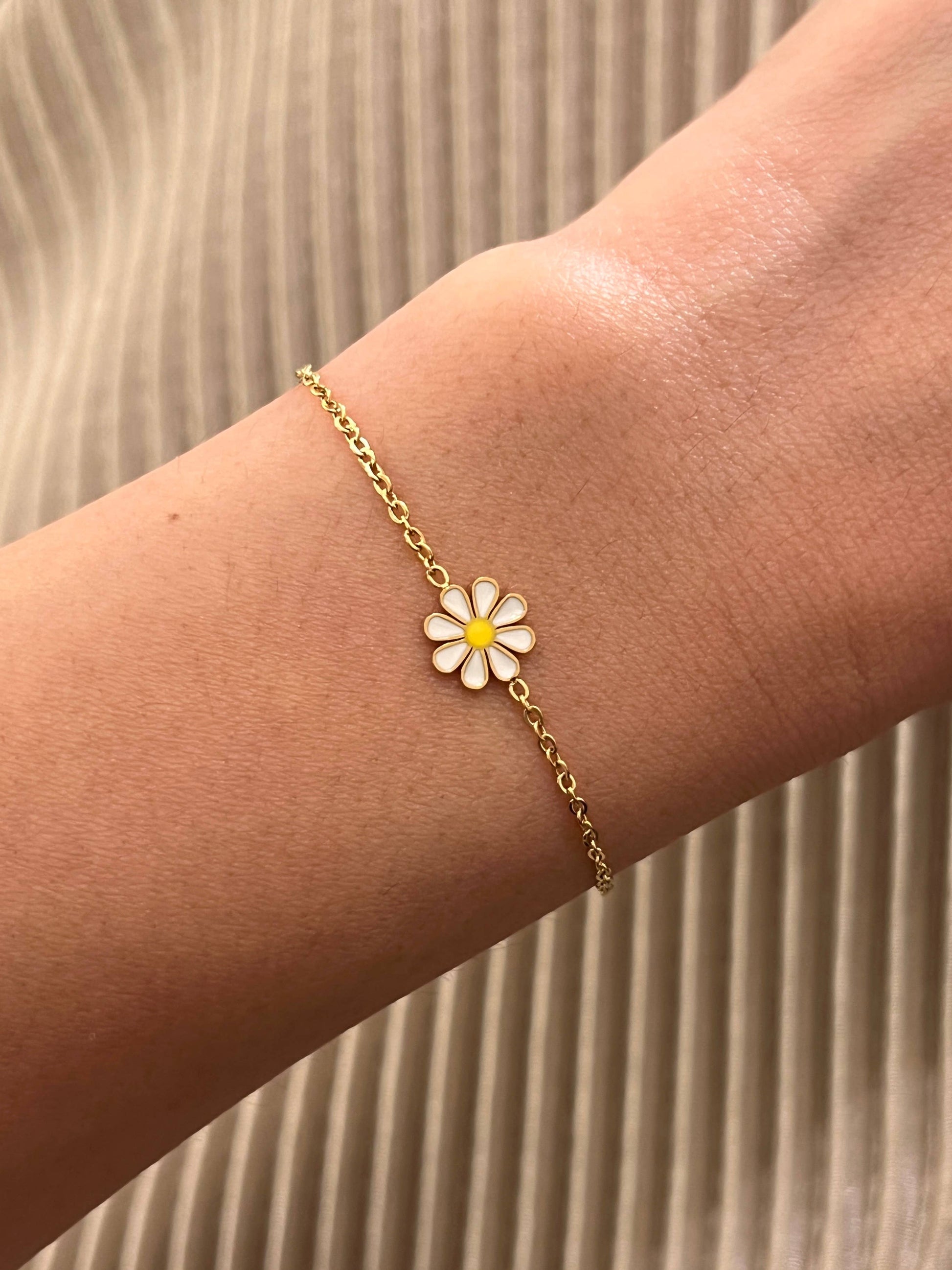 Bracelet fin en acier inoxydable doré composé d'une fine chaîne en maille forçat et d'une petite fleur marguerite aux pétales émaillés blancs et cœur jaune, porté au poignet sur fond de tissu plissé beige.