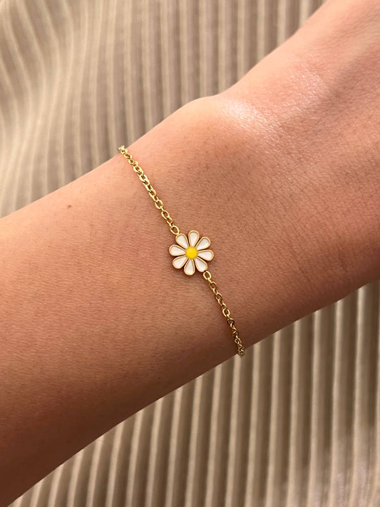 Bracelet fin en acier inoxydable doré composé d'une fine chaîne en maille forçat et d'une petite fleur marguerite aux pétales émaillés blancs et cœur jaune, porté au poignet sur fond de tissu plissé beige.