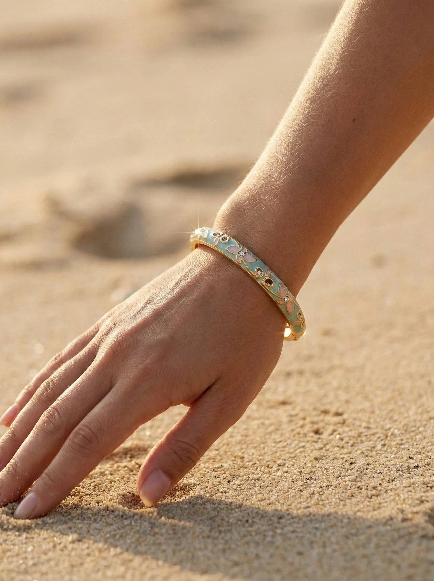 Vue rapprochée d'un bracelet jonc rigide en laiton doré porté au poignet, fond sable chaud. Le bijou présente un émaillage bleu turquoise clair avec de petites fleurs roses et des strass étincelants.