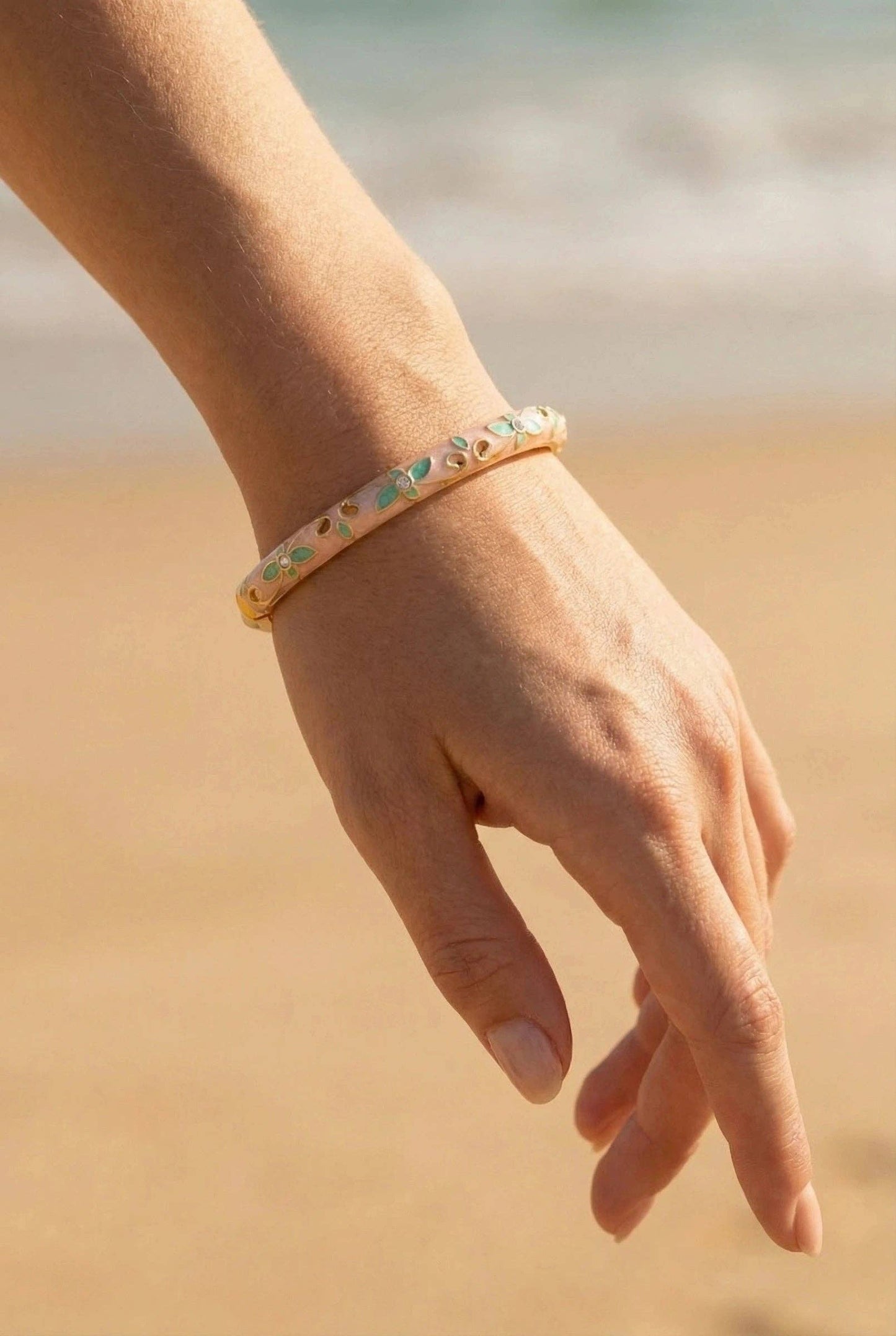 Bracelet rigide type jonc en laiton doré et émail couleur pêche saumonée, orné de motifs floraux vert d'eau et de cristaux clairs, présenté au poignet sur un fond de plage ensoleillée.