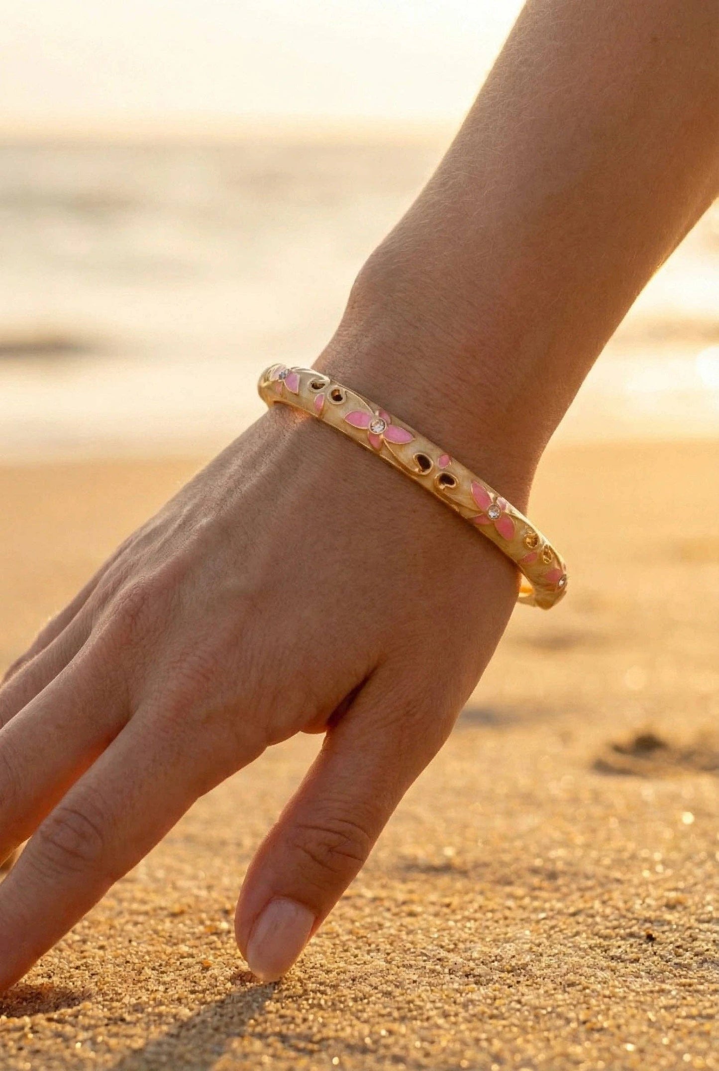 Bracelet jonc rigide ouvrant en laiton doré porté au poignet sur une plage. Le jonc est décoré d'un émail de fond crème, de motifs de fleurs roses et de petits cristaux scintillants au centre.