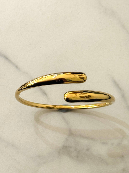 Bracelet rigide de type jonc ouvert et croisé en acier inoxydable doré, doté d'extrémités lisses et bombées en forme de gouttes d'eau au fini miroir très brillant, présenté sur une surface en marbre blanc.