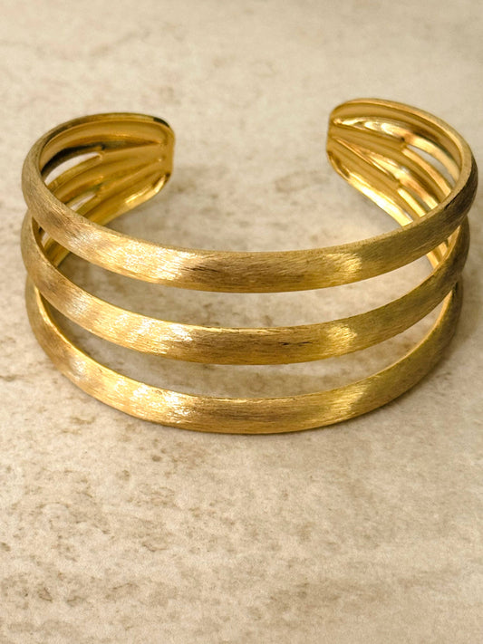 Bracelet jonc rigide de type manchette en acier inoxydable doré, composé de trois rangs parallèles ajourés avec une finition texturée en métal brossé mat, posé sur un fond en pierre beige texturé.