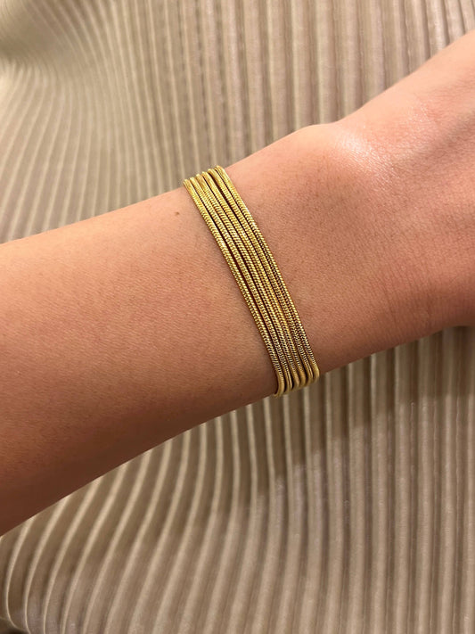 Bracelet large de type manchette composé de cinq rangs de chaînes fines en maille serpent en acier inoxydable doré, porté au poignet, sur un fond de tissu plissé beige doré satiné.