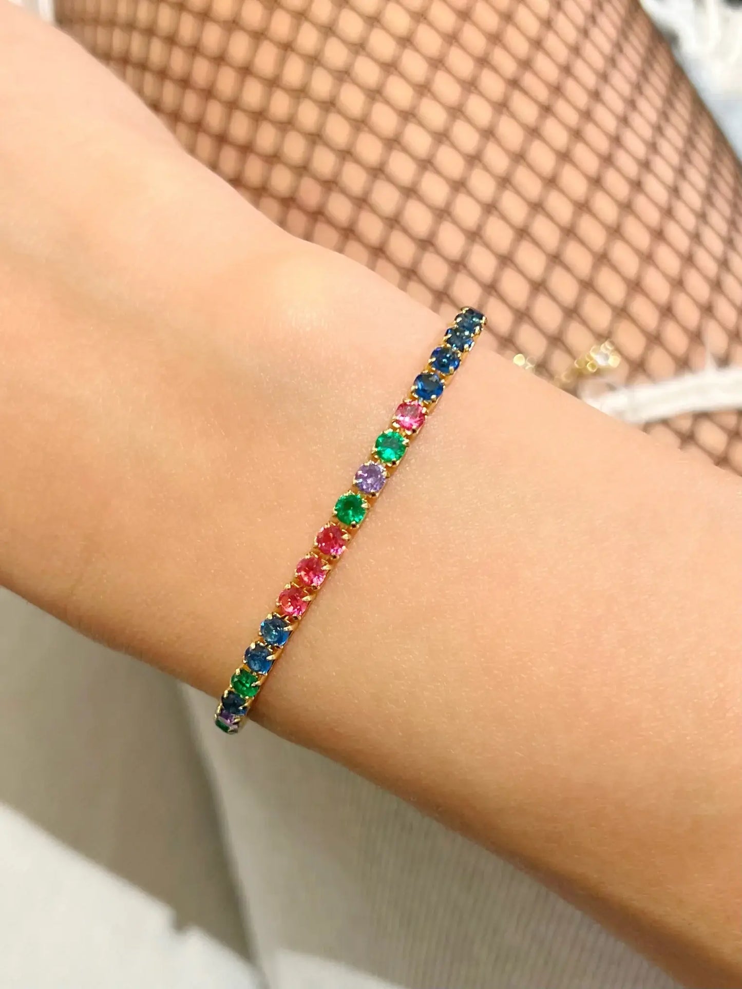 Bracelet de style tennis en acier doré orné d'une suite de petits cristaux ronds multicolores alternant le bleu, le vert, le violet et le rose, porté au poignet.