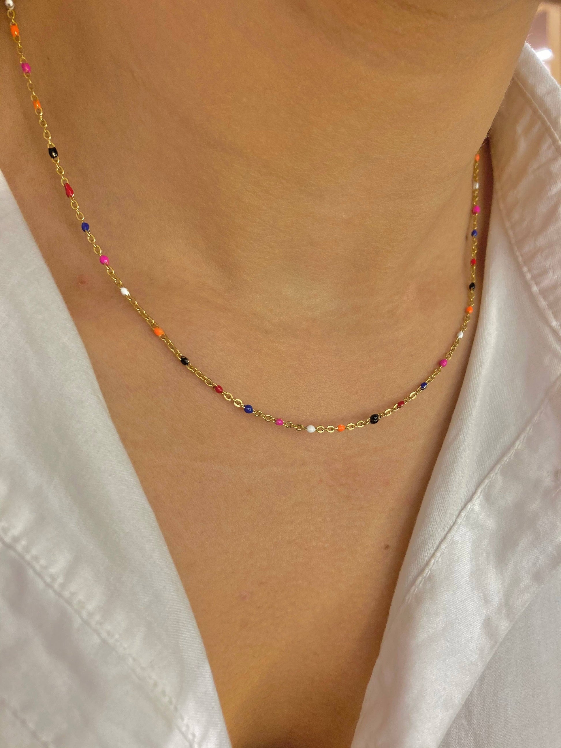 Collier à fine chaîne dorée en acier inoxydable parsemé de petites touches de résine d'émail multicolores (bleu, blanc, noir, orange, rose, rouge), porté autour du cou d'une femme portant une chemise blanche ouverte.
