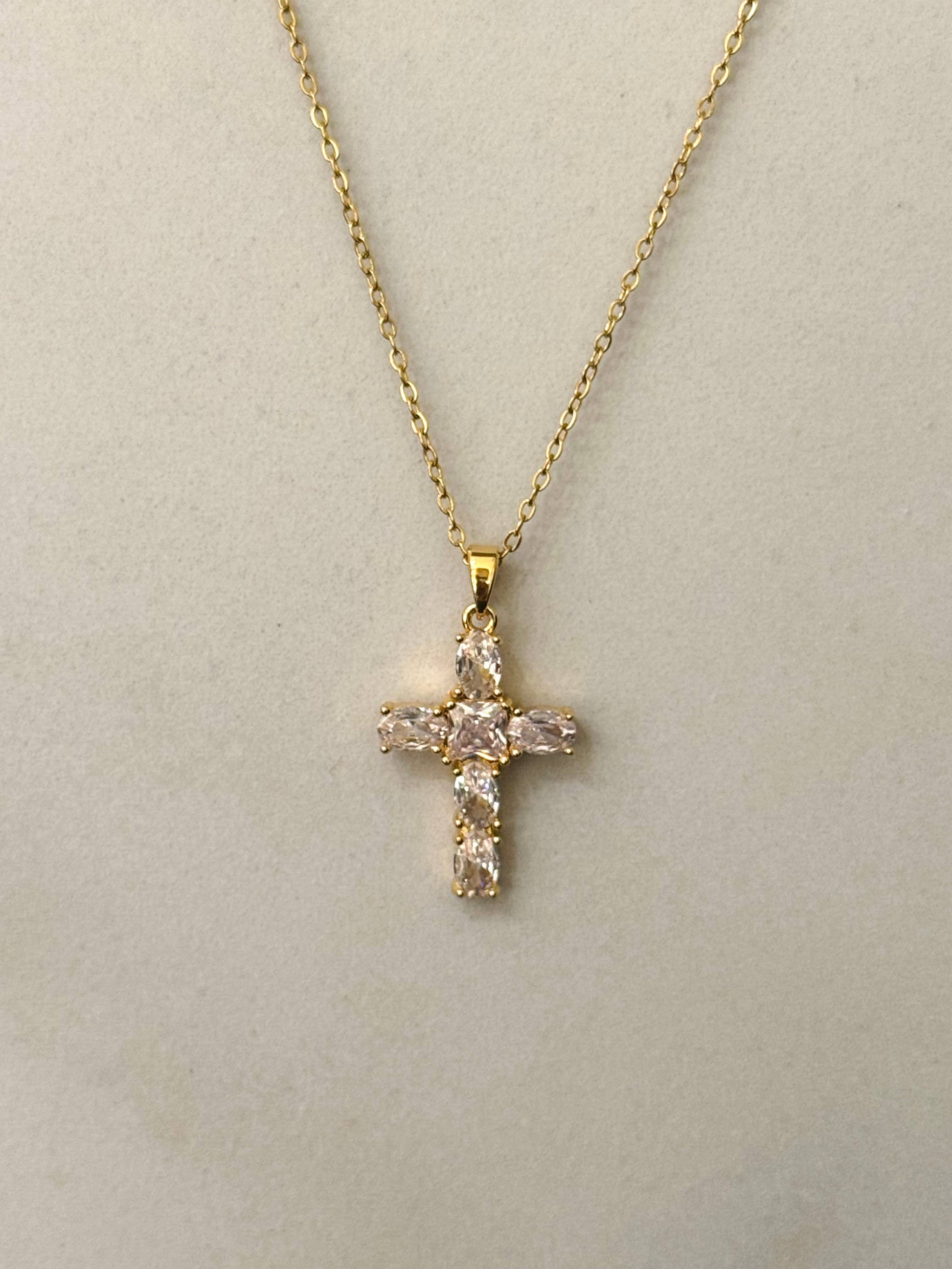 Collier à fine chaîne dorée en acier inoxydable avec un pendentif en forme de croix sertie d'oxydes de zirconium transparents et scintillants, présenté de face sur un fond beige clair épuré.