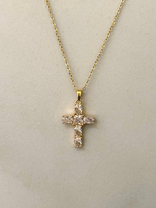 Collier à fine chaîne dorée en acier inoxydable avec un pendentif en forme de croix sertie d'oxydes de zirconium transparents et scintillants, présenté de face sur un fond beige clair épuré.