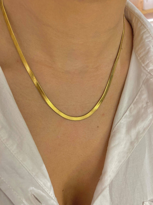 Collier ras-du-cou en acier inoxydable doré formé d'une chaîne à maille serpent plate et lisse très brillante, posée à plat autour du cou d'une femme vêtue d'une chemise en satin blanc ouverte.